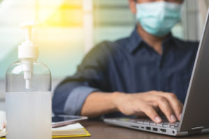 Man wearing a mask on the computer with hand sanitizer nearby