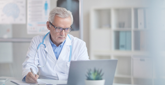 Male doctor in a white coat sitting on his desk in his office working on a laptop