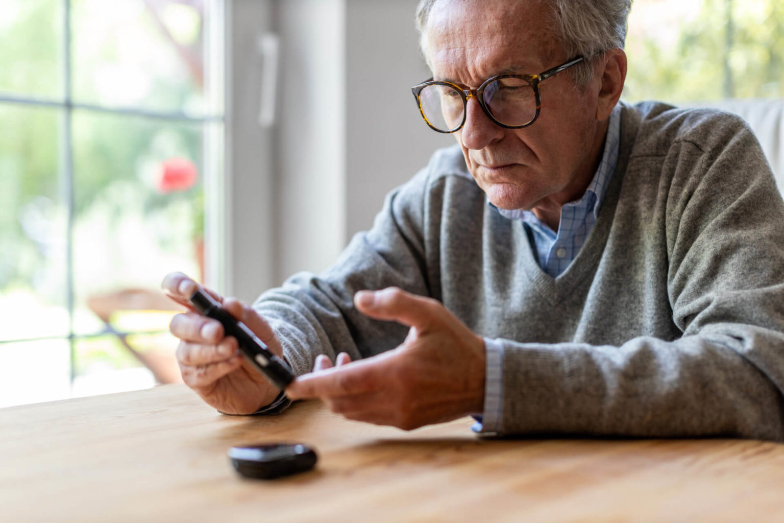 Older man with glasses sitting at a table and checking his blood sugar.